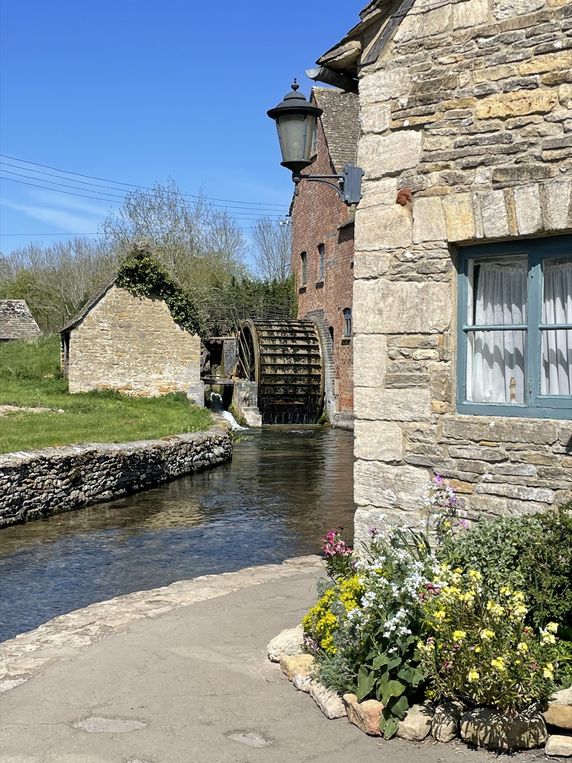 A stone watermill with a waterwheel and flowers beside a stream in the Cotswolds